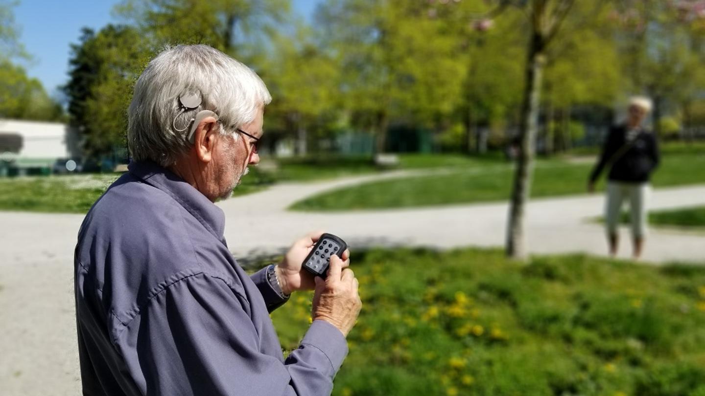 A man stands outside holding a device in his hand with the side of head facing the camera showing his cochlear implant