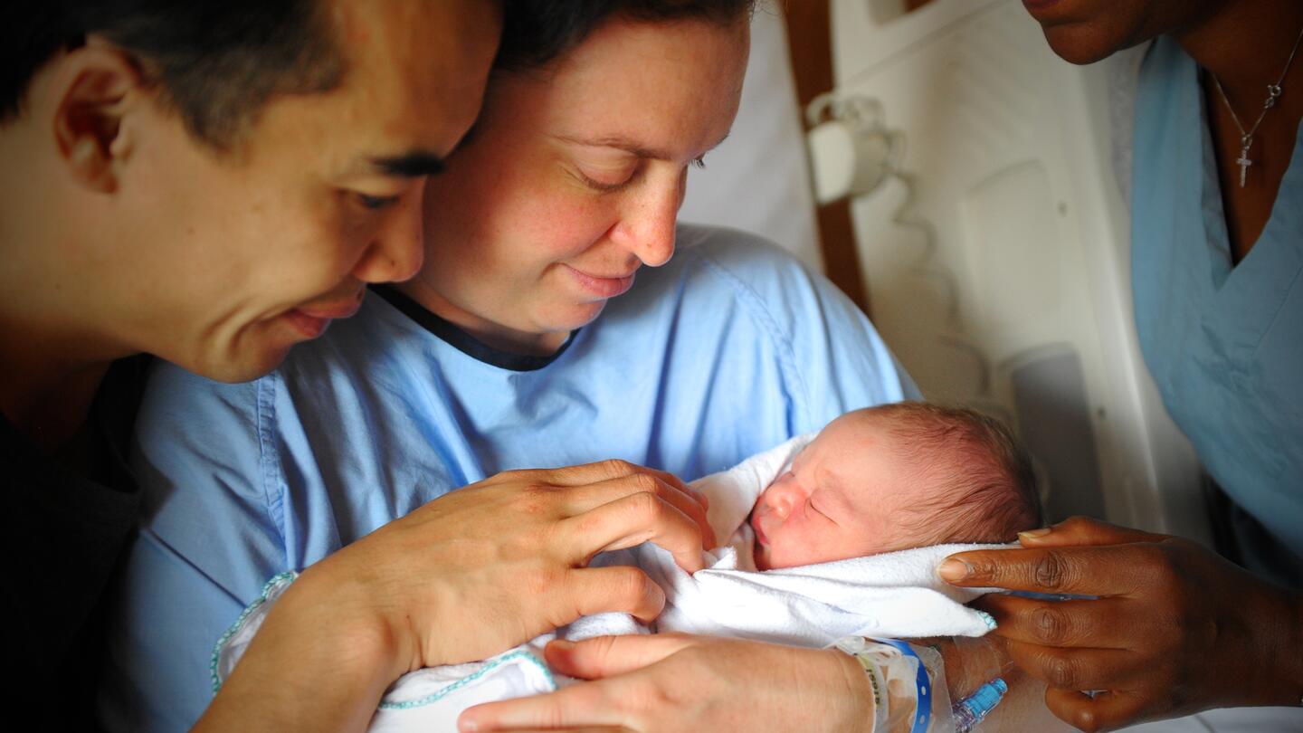 A woman sitting up in a hospital bed with a man standing next to the bed smile down at a newborn baby