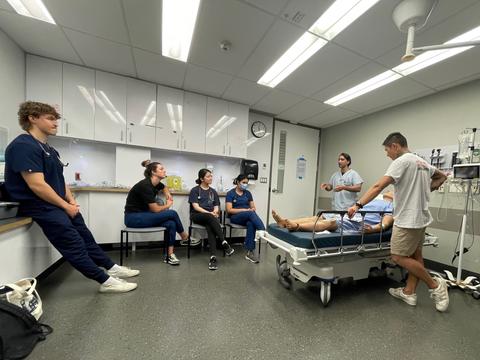 A group of people in scrubs sit and stand around a hospital bed listening to someone speak