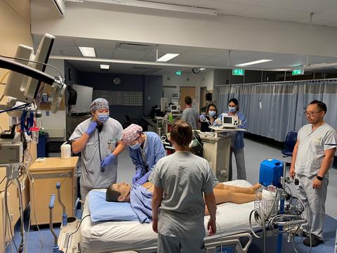 A group of people in scrubs stand around a hospital bed simulating medical care on a mannequin