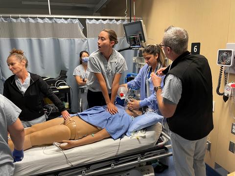 A group of people in scrubs stand around a hospital bed simulating medical care on a mannequin